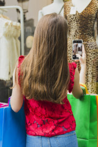 girl in clothing store holding bags taking photo on cell phone of mannequin wearing dress