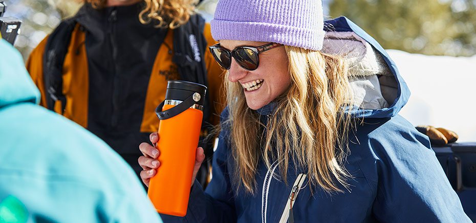 Woman in the snow laughing and holding orange Hydro Flask bottle.