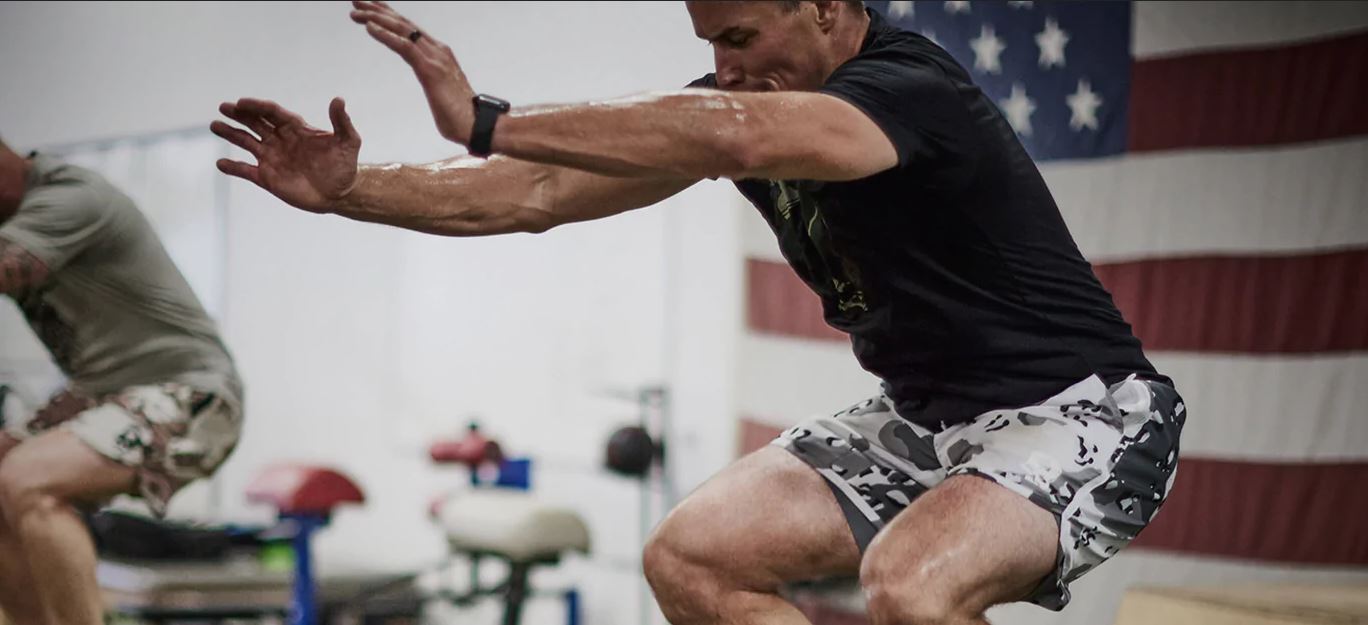 Gentleman wearing shorts and a black shirt in a gym jumping in a squat position with an American flag hanging on the wall behind him.