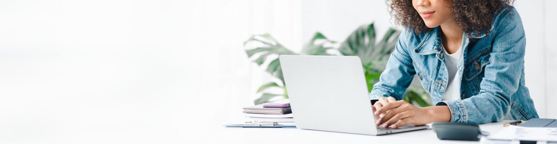 Woman wearing jean jacket typing on a laptop with a plant in the background.