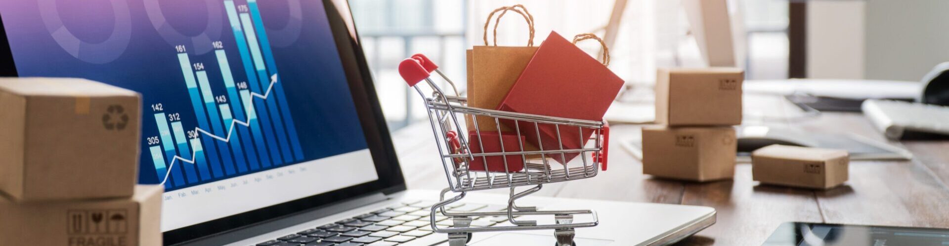 Shopping bags in shopping cart and credit card on laptop with paper boxes on table and sales data economic growth graph on screen, buying and selling services online on network, online shopping and e-commerce concept