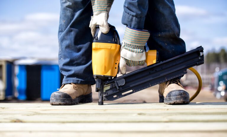 Close up of legs and feet in steel toe boots and arms of man in overalls and work gloves using an air nailer on wood boards outdoors in summer.