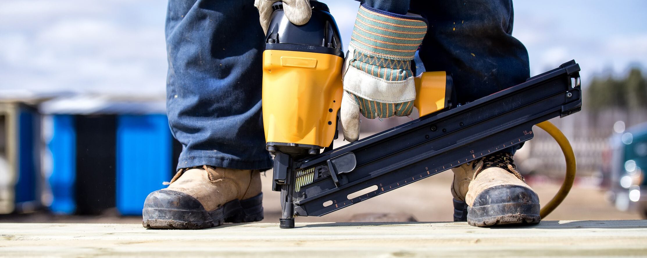 Close up of legs and feet in steel toe boots and arms of man in overalls and work gloves using an air nailer on wood boards outdoors in summer.