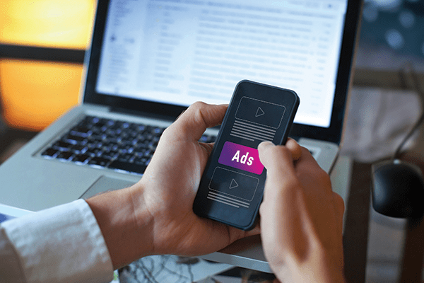 Person using cell phone showing an ad with laptop in the background.