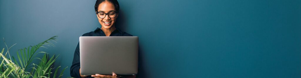 Woman holding laptop and smiling and typing against blue wall next to plant.