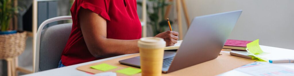 Woman in red shirt writing and typing on a laptop with coffee next to it.