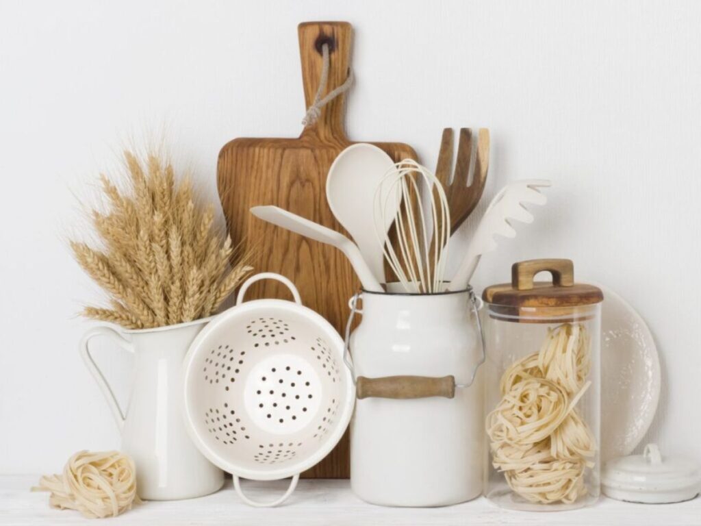 Wooden and white kitchen utensils with a cutting board, colander and jar of pasta beside dried wheat