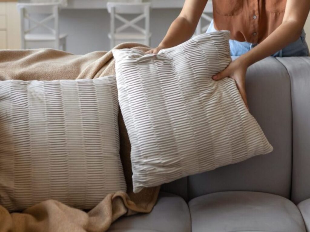 Person arranging textured cream throw pillows on a gray sofa with a tan blanket