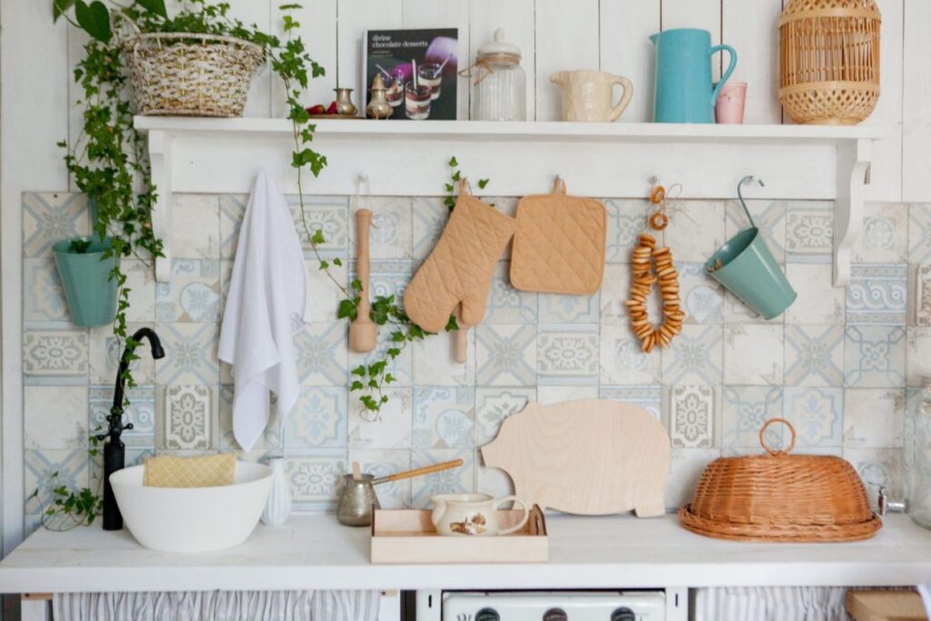 White kitchen shelf above a blue and white patterned tile backsplash, with hanging oven mitts, a towel and trailing ivy