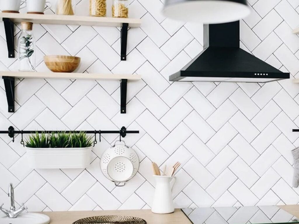 White herringbone tile kitchen backsplash with open shelves, a black range hood and a hanging colander